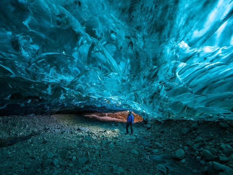 A vast turquoise ice cave roof over a dark gravel floor, one visitor small beneath it