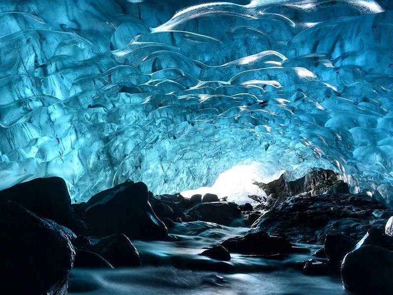 The scalloped blue ceiling of an ice cave over black rocks and running meltwater