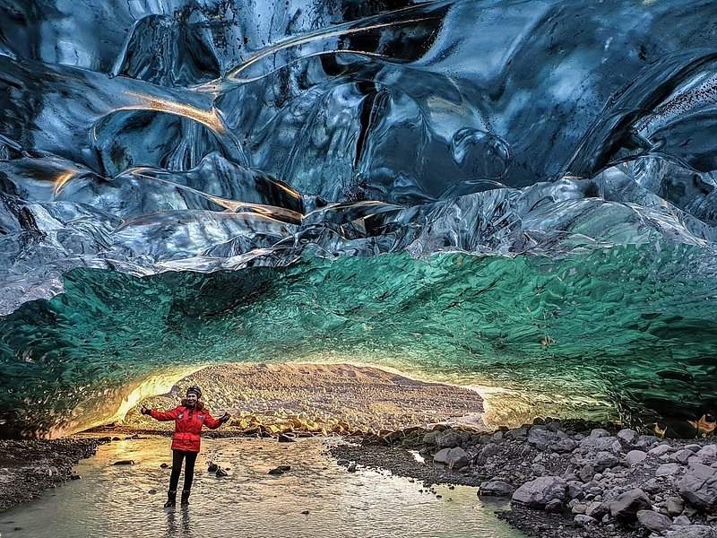 A dark ice cave mouth glowing green and gold, one visitor small on the gravel