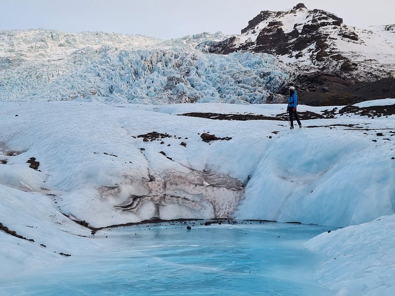 A lone hiker on a crevassed outlet glacier above a turquoise meltwater pool
