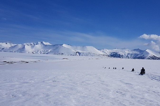 A line of snowmobiles crossing a flat white ice cap below snow-covered peaks