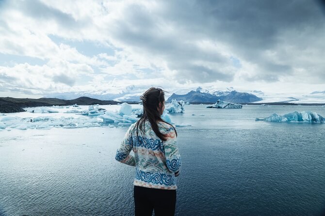 A traveller seen from behind on the shore of an iceberg lagoon under heavy cloud